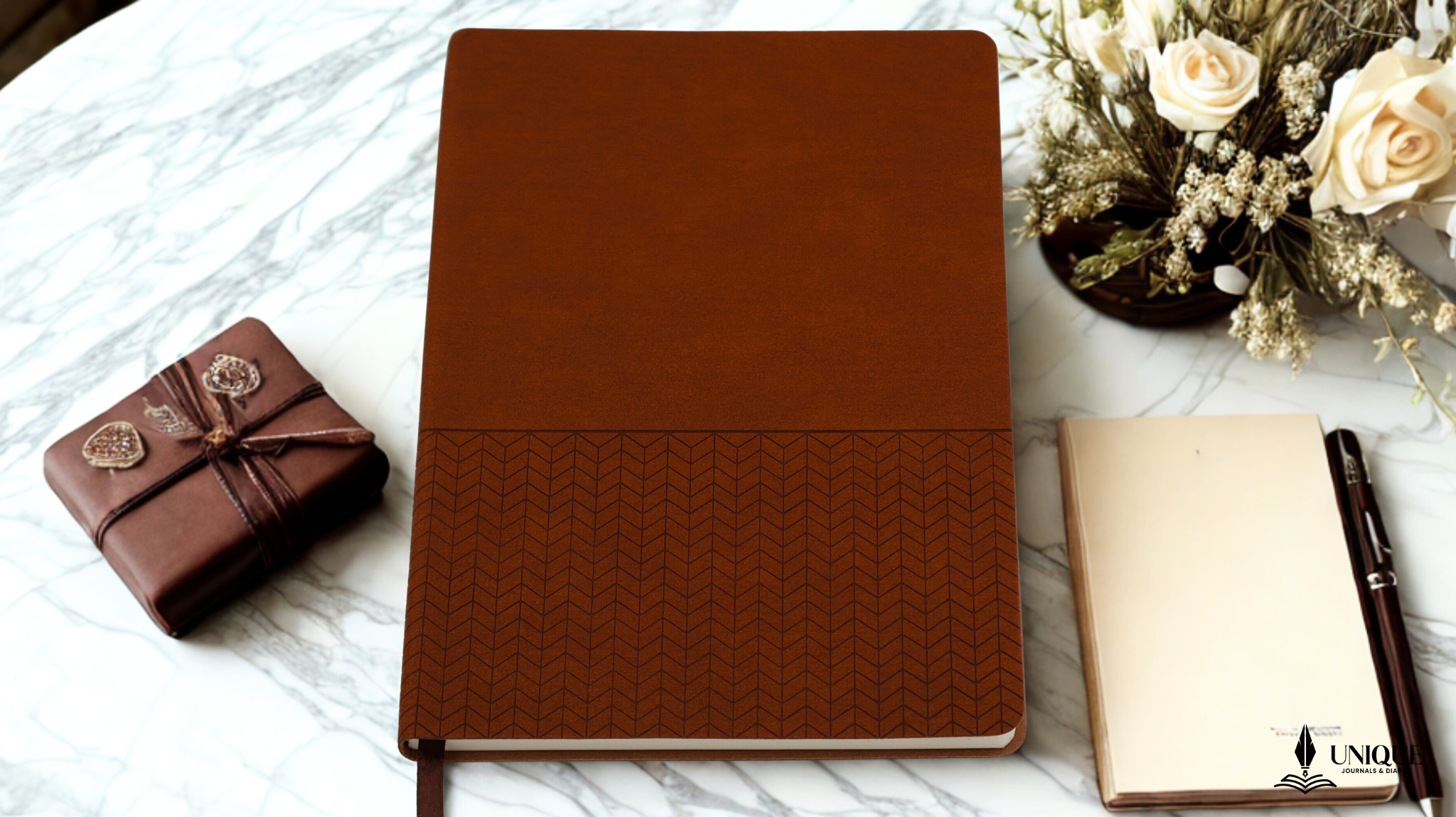 Brown leather notebook on a marble surface with a small gift box and flowers.


